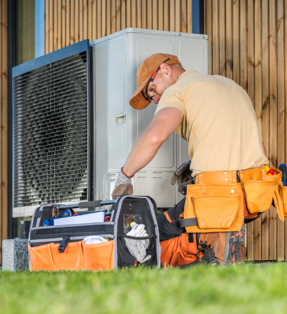 A technician in an orange cap and tool belt works on an outdoor air conditioning unit, with a toolbag beside him on the grass.
