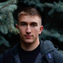 A man with short hair, wearing a dark jacket, stands outdoors near pine tree branches, looking at the camera.