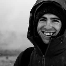 A person wearing a hooded jacket and beanie smiles in a snowy outdoor setting. Black and white image.