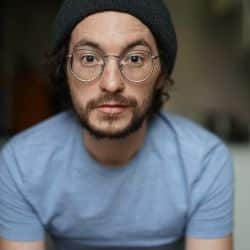 Man with glasses and a beard wearing a black beanie and light blue shirt, facing the camera.